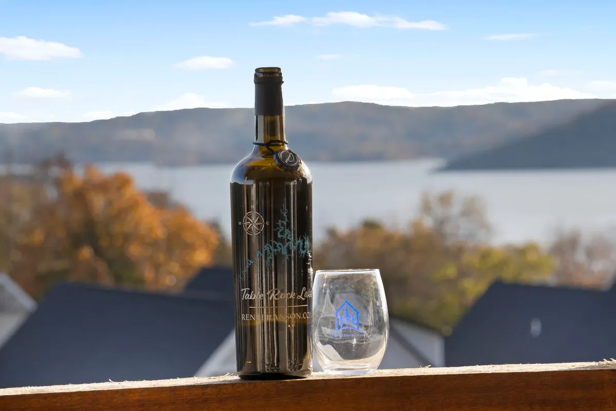 A tranquil outdoor scene featuring a wine bottle and glass on a table, with the distant lake shimmering below. The composition highlights the enjoyment of drinks while admiring the sunset over the water from the deck.