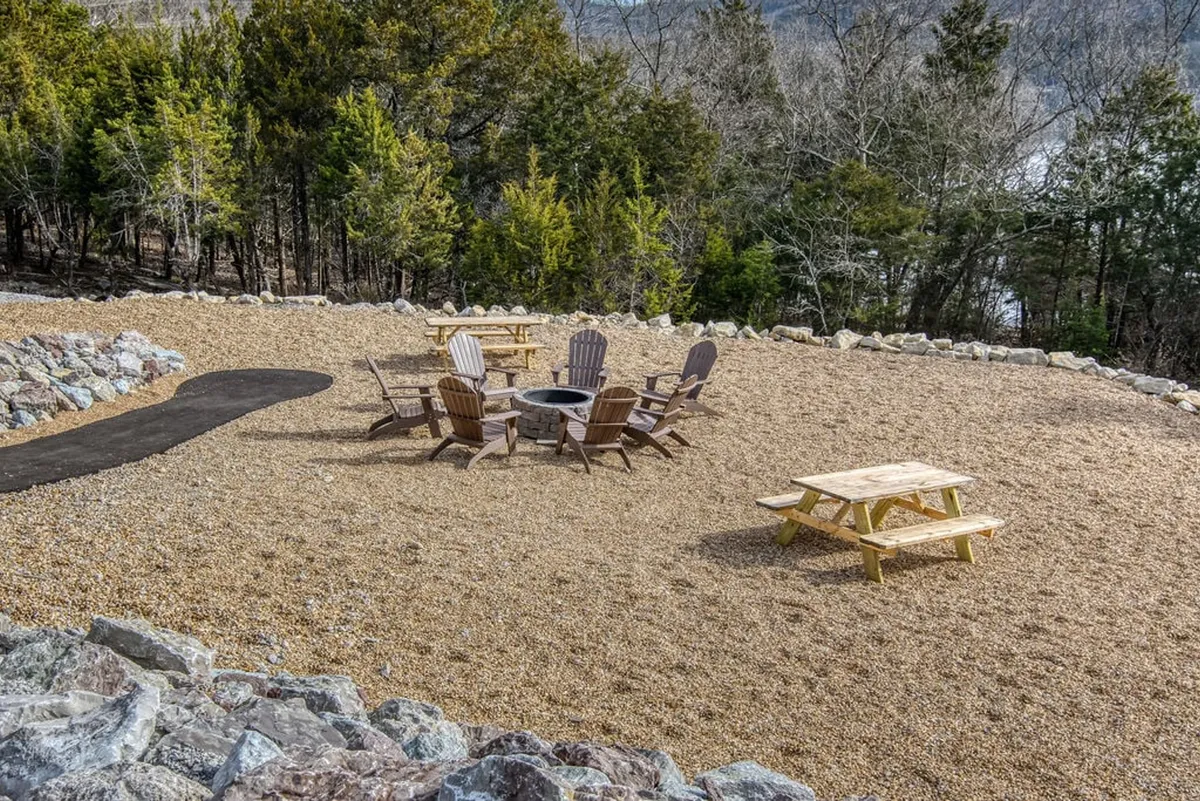 A welcoming outdoor communal firepit surrounded by adirondack chairs and a picnic table, set within a landscaped stone circle. The setting is perfect for evening gatherings, storytelling, and making memories under the stars.