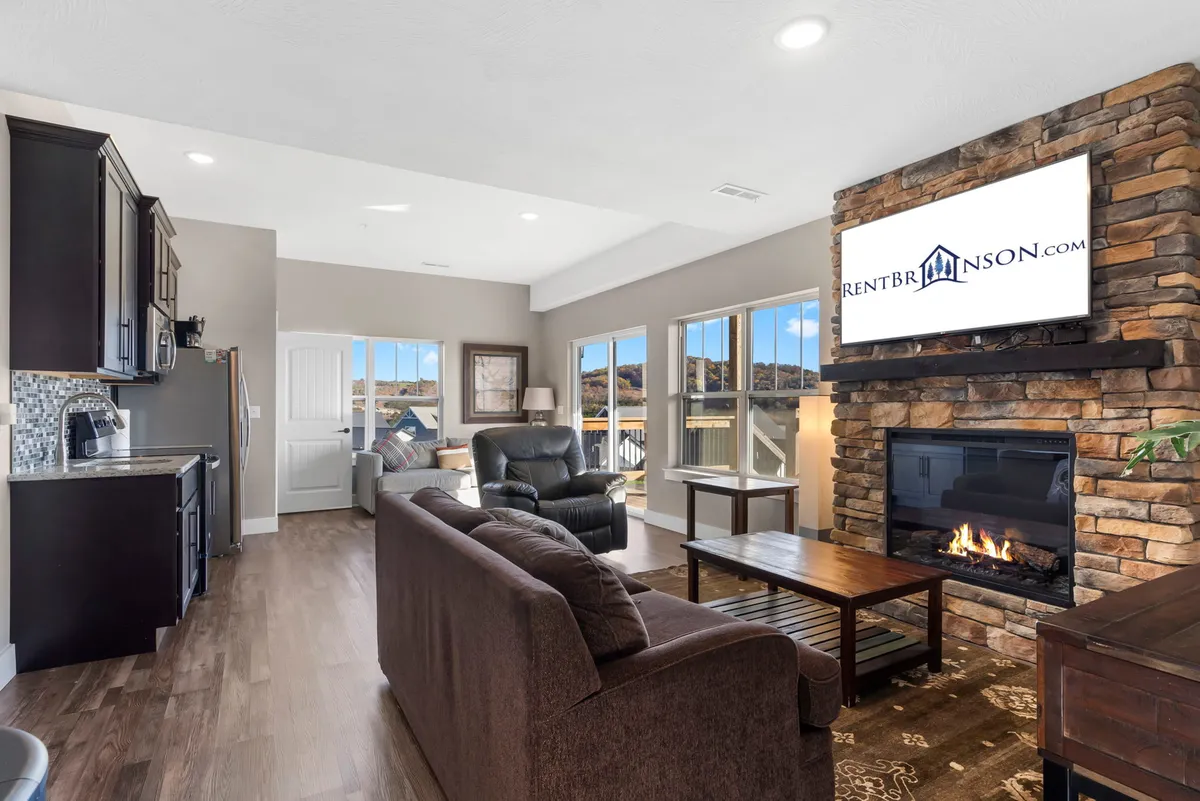 A cozy living space with a stone fireplace, comfortable sofas, a flat-screen TV, and large windows that let in natural light. The kitchen area is visible in the background, tying the living space to the heart of the home.