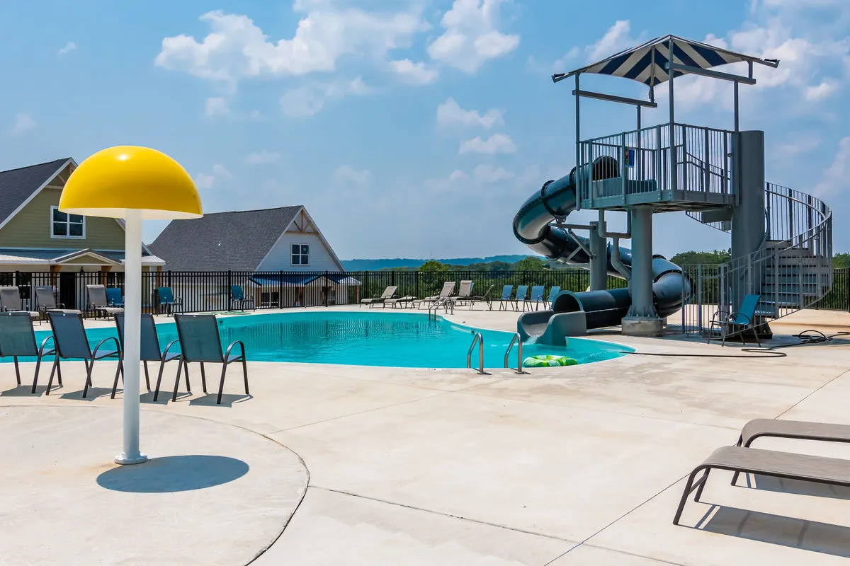 An exterior view of the outdoor swimming pool area, featuring a fun water slide, a bright yellow umbrella, and several lounge chairs. The pool is surrounded by landscaped greenery, providing a lively and relaxing atmosphere ideal for group activities and summer fun.