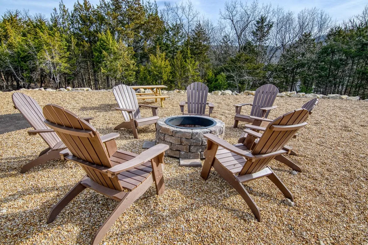 A cozy communal fire pit surrounded by Adirondack chairs, nestled among trees and gravel ground. The setting evokes warm evenings spent sharing stories and making s'mores under the stars.
