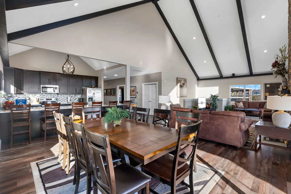 This image captures an open floor plan featuring a vaulted ceiling, a welcoming living room with a fireplace, a visible kitchen with a central island, a dining table, and large windows that bring in abundant natural light.