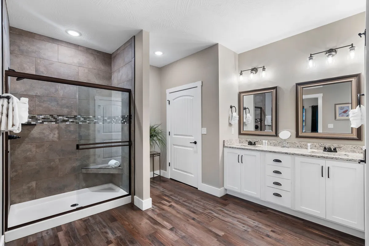 A luxurious en-suite bathroom featuring a glass shower enclosure, double vanity with mirrors, granite countertop, and warm wooden flooring. The space feels serene and upscale.