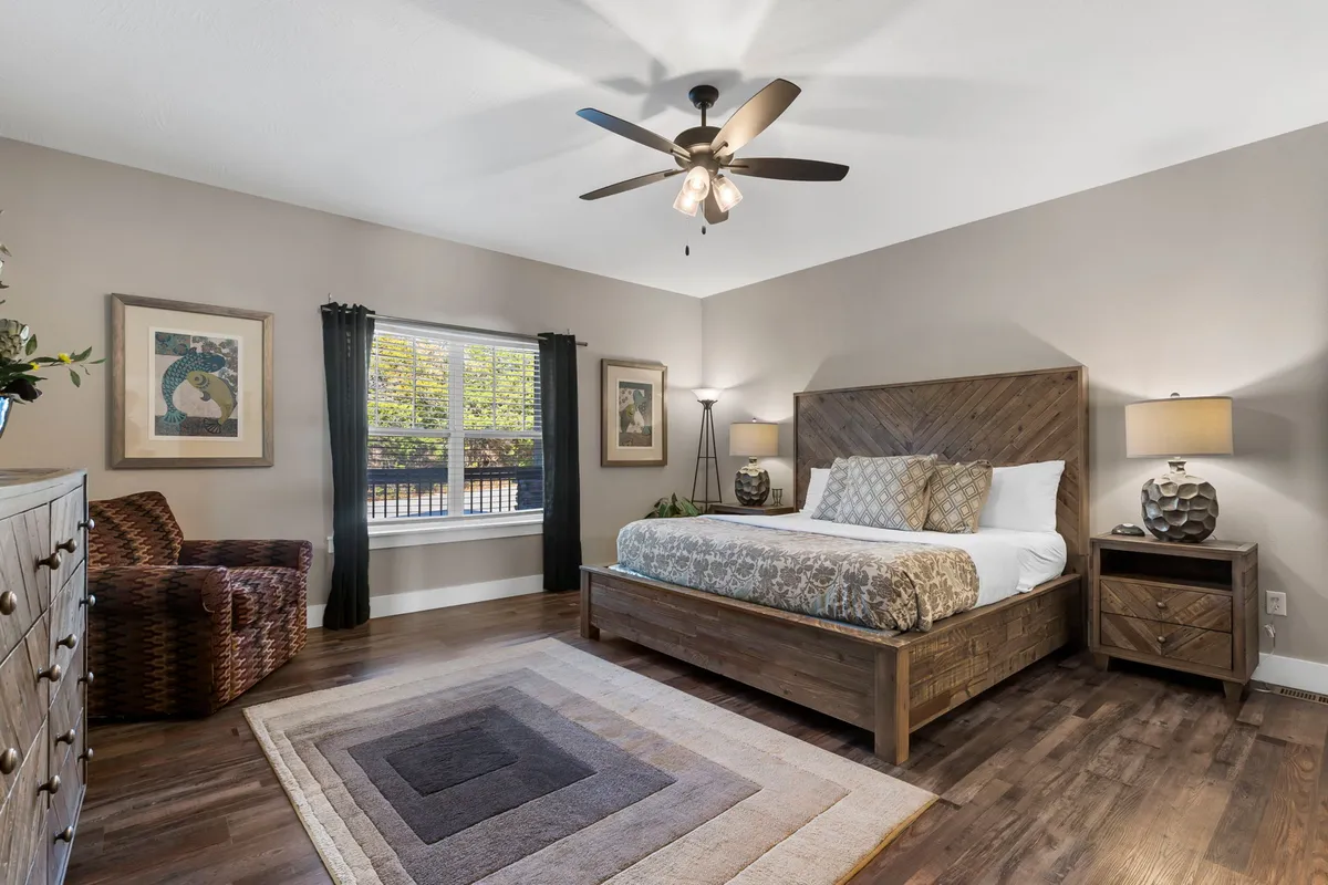 A serene, spacious bedroom featuring a plush bed flanked by nightstands and an armchair by the window. Natural light fills the room, accentuating calming tones and inviting textures.